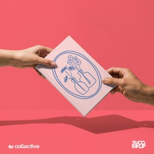 May include: A light pink hardcover book featuring a dark blue line art illustration of two vases containing flowers inside an oval frame. The book is held at an angle against a solid pink background. The text S/ collective and SVOO MKUP appear.