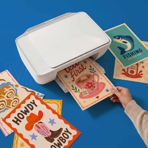 May include: A white printer printing colorful cards with various designs. One card features a teacup with the text "Tea First". Other cards show a fish, a cowboy boot, and a tomato sauce graphic. The background is a solid blue.