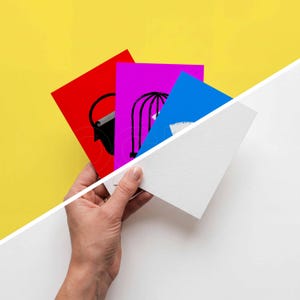K&ouml;nnte beinhalten: Eine Hand h&auml;lt einen Stapel bunter Karten vor einem gelben und wei&szlig;en Hintergrund. Die Karten sind rot, lila und blau, jede mit einer schwarzen Strichzeichnung. Die rote Karte hat einen Eimer, die lila Karte einen K&auml;fig und die blaue Karte eine wei&szlig;e Form.