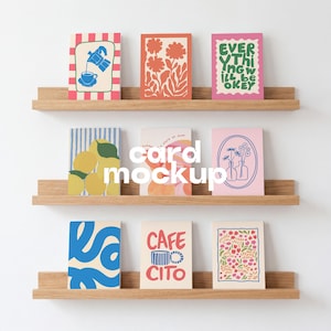 May include: Three light wood floating shelves displaying nine rectangular greeting cards with various colorful illustrations, including a coffee pot, flowers, lemons, and text reading "EVERYTHING WILL BE OKEY" and "CAFE CITO" on a plain white wall.