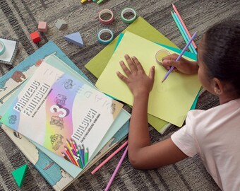 May include: A young person is drawing on a yellow sheet of paper with a purple pencil. The paper is on top of a green and blue folder. There are several colorful pencils and rolls of tape on the floor. The person is using their left hand to hold the paper in place. The person is wearing a pink shirt.