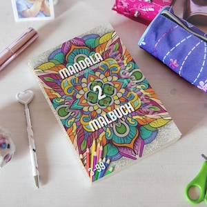 Op de afbeelding: Een kleurrijk mandala kleurboek met de tekst "MANDALA" en "MALBUCH" is omringd door andere schrijfwaren. Het boek heeft een levendig bloemendesign met het nummer "2" in het midden. Een pen met hartvormige top en een roze etui zijn ook zichtbaar.