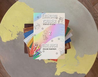 Puede incluir: Un libro colorido con un fondo de arcoíris y estrellas. El título es "Einhorn Zauber" y el subtítulo es "Die bunte Welt der Fantasie". El libro es un libro para colorear para niños y tiene el número "2" en la portada. El libro está encima de una pila de otros libros.