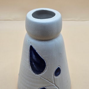 May include: A light gray ceramic vase with a flared base and a rounded top. The vase features a dark blue leaf design. The vase is approximately 5 inches tall.