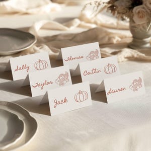 May include: White place cards with red lettering and illustrations. Each card displays a name, along with a pumpkin or turkey illustration. The cards are arranged on a white tablecloth, suggesting a Thanksgiving or autumn theme.