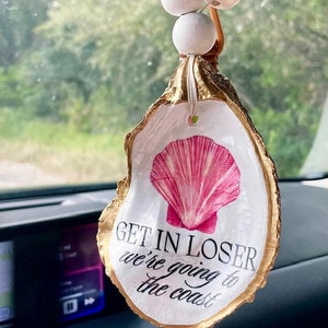 May include: A white oyster shell car charm with gold accents. The shell has a pink seashell image and the text "Get in loser we're going to the coast".