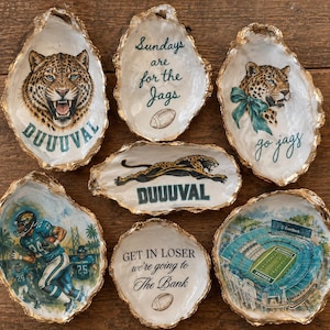 May include: Seven decorative oyster shells with gold-painted edges, featuring Jacksonville Jaguars-themed illustrations including a jaguar head, a football stadium, a player, and text such as DUUUVAL and Sundays are for the Jags.
