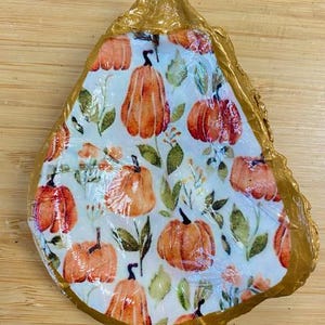 May include: A decorative oyster shell with a gold-painted edge. The shell's interior features a pumpkin-themed design with orange pumpkins, green leaves, and small orange flowers on a white background. The shell is placed on a wooden surface.