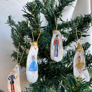 May include: Four hand-painted oyster shell ornaments featuring characters from the Nutcracker ballet. The ornaments are decorated with gold paint and feature a nutcracker, a ballerina in a blue dress, a ballerina in a gold dress, and a king.