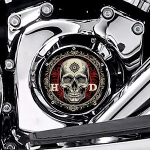 Puede incluir: Una cubierta de motor de motocicleta con un diseño de calavera negra y roja. La calavera está centrada con un diseño floral en la frente, rodeada por un patrón floral rojo y las letras "H D". La cubierta está sobre un fondo cromado brillante.