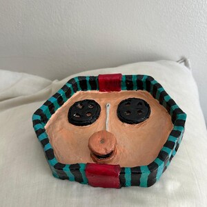 May include: A handmade, hexagonal decorative tray with a face design. The tray features a peach-colored face with black button eyes and a button nose. The tray's border is black with teal stripes and red accents. The tray is sitting on a white surface.