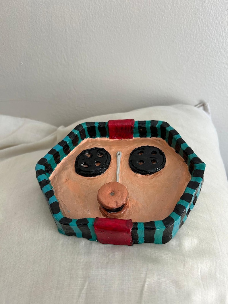 May include: A handmade, hexagonal decorative tray with a face design. The tray features a peach-colored face with black button eyes and a button nose. The tray's border is black with teal stripes and red accents. The tray is sitting on a white surface.