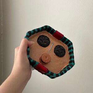 May include: A handmade, decorative tray with a tan interior, black buttons, and a spool of thread. The tray's border is black and teal striped, with red accents. A needle is placed between the buttons.