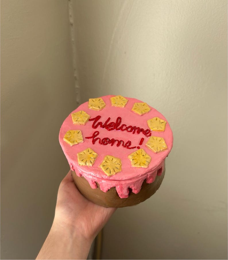 Puede incluir: Una caja redonda y decorativa que se asemeja a un pastel, sostenida por una mano. La parte superior es rosa con formas de pi&ntilde;a amarillas y texto rojo que dice "Welcome home!". Los lados son marrones con glaseado rosa.