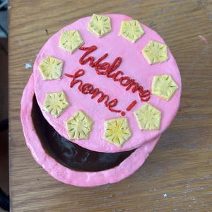 Puede incluir: Una caja de baratijas redonda y rosa con tapa. La tapa est&aacute; decorada con dise&ntilde;os amarillos en forma de estrella y las palabras "welcome home" en escritura roja. La caja est&aacute; abierta, revelando un interior oscuro.