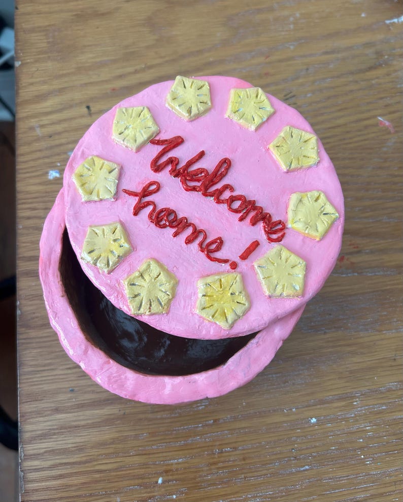 Puede incluir: Una caja de baratijas redonda y rosa con tapa. La tapa est&aacute; decorada con dise&ntilde;os amarillos en forma de estrella y las palabras "welcome home" en escritura roja. La caja est&aacute; abierta, revelando un interior oscuro.