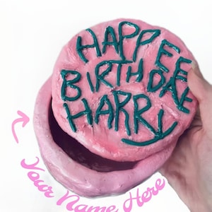May include: Pink ceramic box with a lid that has the words "Happy Birthday Harry" written on it in green. The box is open and the lid is being held by a hand. The words "Your Name Here" are written on the bottom of the box.