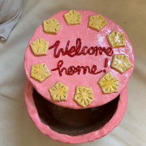 Puede incluir: Una caja de baratijas redonda rosa con tapa. La tapa est&aacute; decorada con formas de pi&ntilde;a amarillas y las palabras "Welcome home!" en rojo. La caja est&aacute; abierta, revelando el interior.
