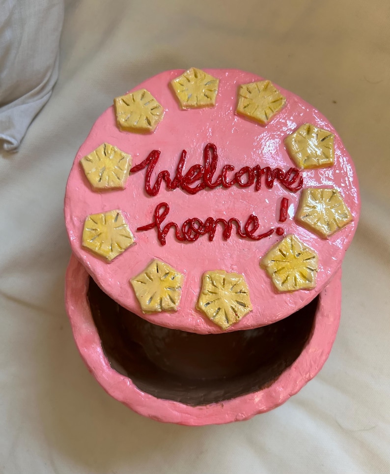 Puede incluir: Una caja de baratijas redonda rosa con tapa. La tapa est&aacute; decorada con formas de pi&ntilde;a amarillas y las palabras "Welcome home!" en rojo. La caja est&aacute; abierta, revelando el interior.