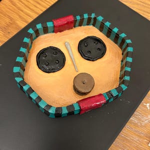 May include: A handmade, painted tray with a face design. The tray features a tan interior with two large black button eyes, a small brown button nose, and a silver needle. The border is teal and black striped with red accents.
