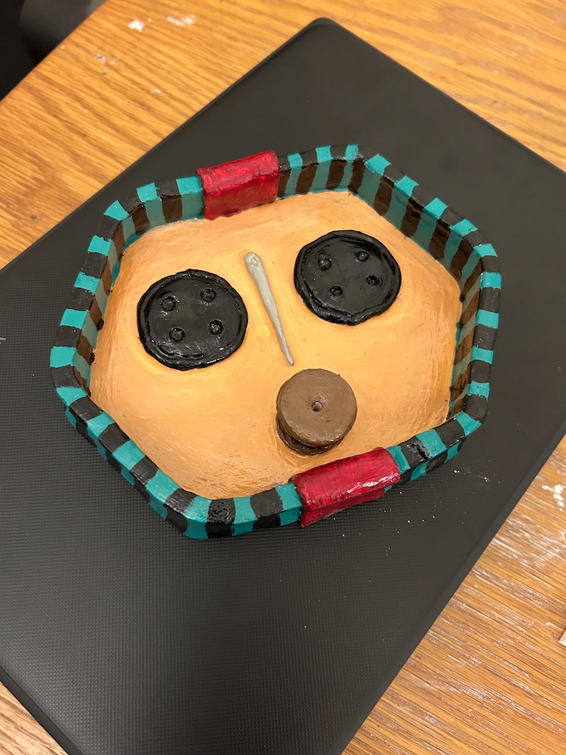 May include: A handmade, painted tray with a face design. The tray features a tan interior with two large black button eyes, a small brown button nose, and a silver needle. The border is teal and black striped with red accents.