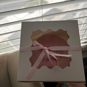 Puede incluir: Una caja de panader&iacute;a blanca con una ventana festoneada que revela un pastel rosa decorado con detalles amarillos y la palabra "name" en cursiva. Una cinta rosa est&aacute; atada alrededor de la caja.