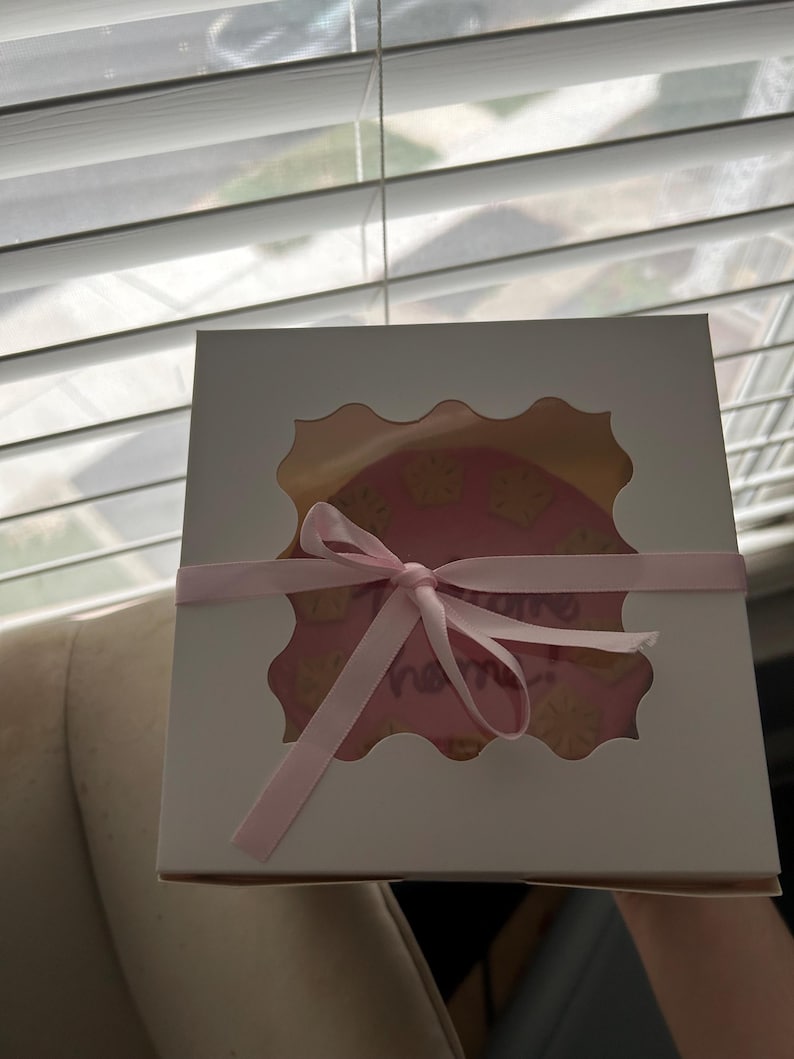 Puede incluir: Una caja de panader&iacute;a blanca con una ventana festoneada que revela un pastel rosa decorado con detalles amarillos y la palabra "name" en cursiva. Una cinta rosa est&aacute; atada alrededor de la caja.