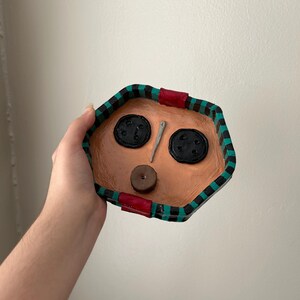 May include: A handmade, octagonal-shaped trinket dish with a tan interior and a black, green, and red patterned border. The dish features two large black button accents, a small brown button, and a silver needle.