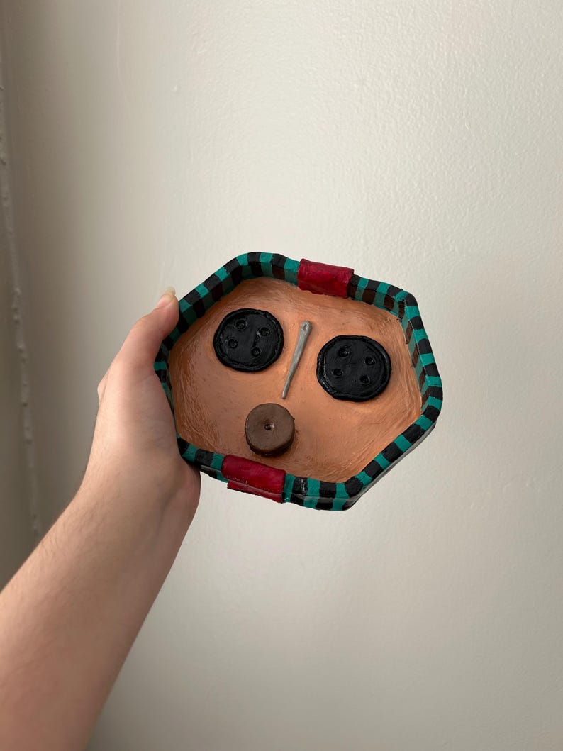 May include: A handmade, octagonal-shaped trinket dish with a tan interior and a black, green, and red patterned border. The dish features two large black button accents, a small brown button, and a silver needle.