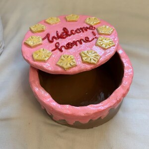 Puede incluir: Una caja redonda y abierta para baratijas con tapa, pintada en tonos rosa y marr&oacute;n. La tapa est&aacute; decorada con acentos amarillos en forma de estrella y las palabras "Welcome home!" en escritura roja. La caja est&aacute; abierta, revelando un interior marr&oacute;n.