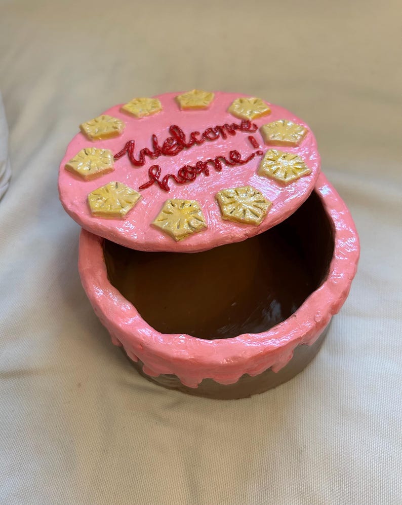 Puede incluir: Una caja redonda y abierta para baratijas con tapa, pintada en tonos rosa y marr&oacute;n. La tapa est&aacute; decorada con acentos amarillos en forma de estrella y las palabras "Welcome home!" en escritura roja. La caja est&aacute; abierta, revelando un interior marr&oacute;n.