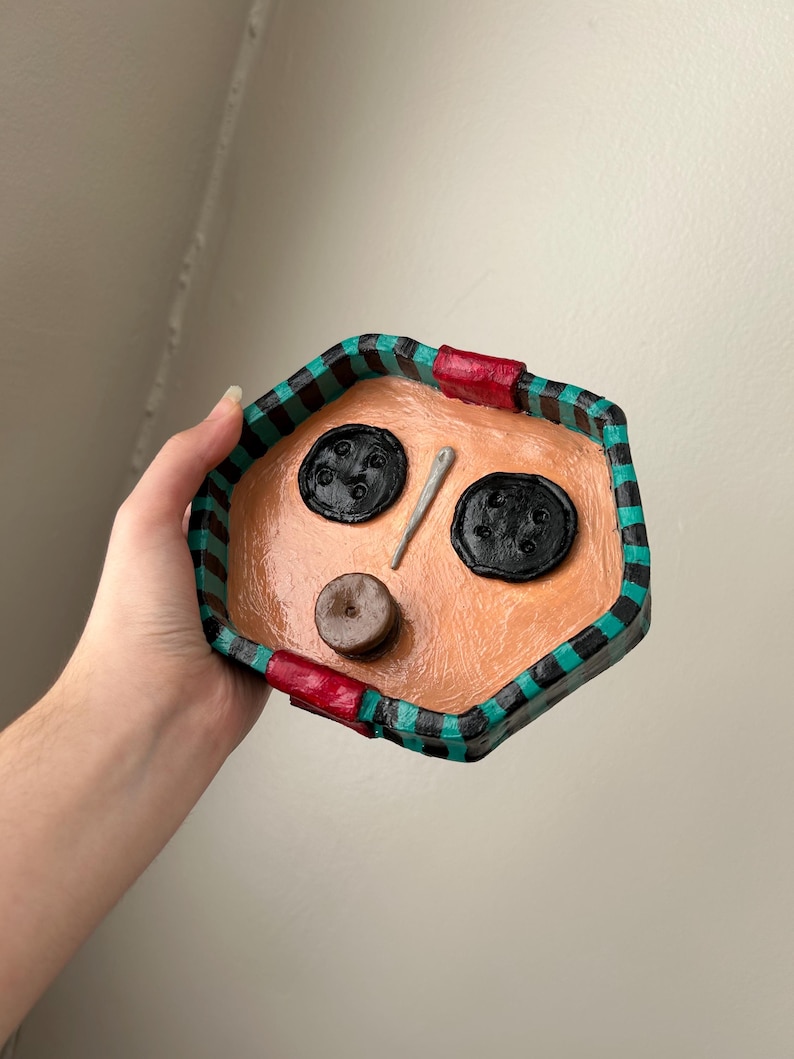 May include: A handmade, hexagonal tray with a peach-colored interior and a black, green, and red plaid border. The tray features two large black buttons, a small brown button, and a silver needle. The tray is held by a hand.