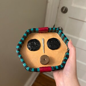 May include: A handmade, hexagonal trinket dish with a face design. The dish features two black button eyes, a needle nose, and a button mouth. The rim is painted with black, teal, and red accents. The dish is tan-colored.