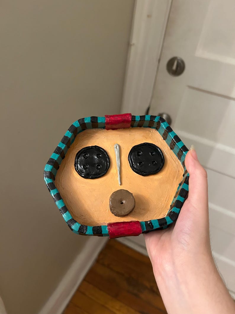 May include: A handmade, hexagonal trinket dish with a face design. The dish features two black button eyes, a needle nose, and a button mouth. The rim is painted with black, teal, and red accents. The dish is tan-colored.