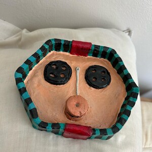 May include: A handmade, hexagonal-shaped clay tray with a face design. The tray features two large black button eyes, a small button nose, and a circular button mouth. The tray's border is black, teal, and red.