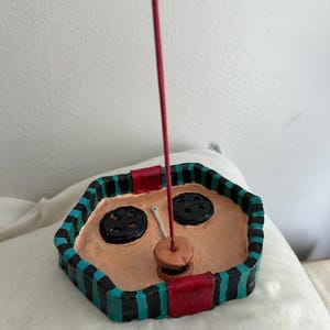 May include: Handmade ceramic incense holder with a unique design. The holder is hexagonal with a tan interior and a black, teal, and red striped border. Features two black button-shaped accents and a red incense stick.