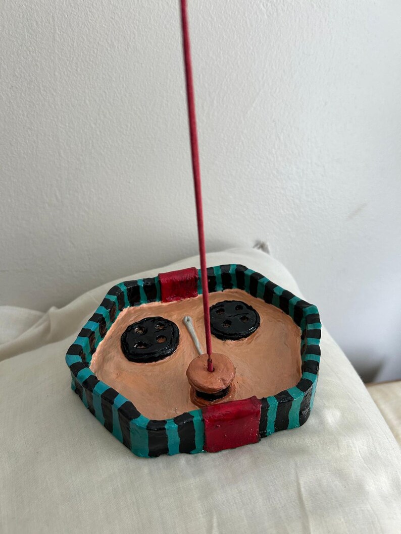 May include: Handmade ceramic incense holder with a unique design. The holder is hexagonal with a tan interior and a black, teal, and red striped border. Features two black button-shaped accents and a red incense stick.