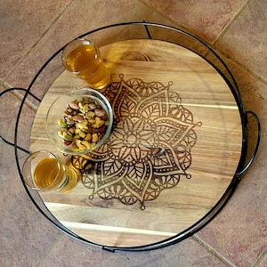 May include: A wooden serving tray with a black metal rim and a decorative mandala design. The tray has three glasses and a bowl of nuts on it.