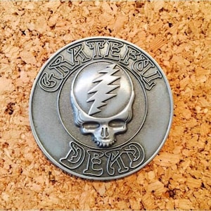 May include: A silver-colored metal emblem featuring the Grateful Dead's Steal Your Face skull and lightning bolt design. The words "Grateful Dead" and "DEK" are inscribed around the skull. The emblem is circular and set against a cork background.