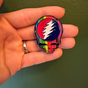 May include: A rainbow-colored enamel pin featuring the Grateful Dead's iconic skull and lightning bolt logo.