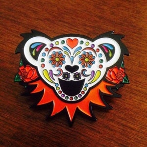 May include: A colorful Day of the Dead-style enamel pin featuring a smiling bear with a sugar skull design. The bear has a red heart, black eyes, and a white background with colorful floral details.