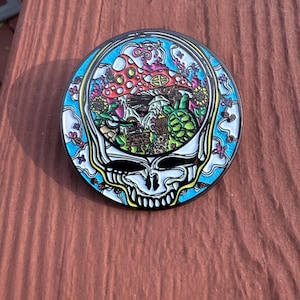 May include: A round, colorful pin featuring a skull design with a psychedelic mushroom and floral motif. The pin has a black border, a blue background, and a vibrant array of colors including red, green, and yellow. The skull has a smiling expression.