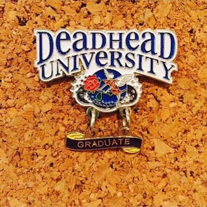 May include: A pin with the words "Deadhead University" in blue and white lettering. The pin features a rose, a hummingbird, and the word "GRADUATE" on a banner. The pin is set against a corkboard background.