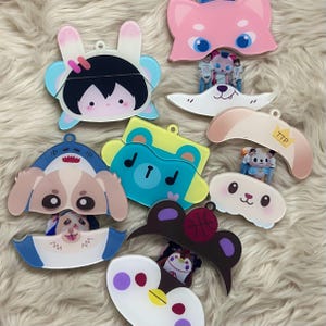 Könnte beinhalten: Eine Sammlung von Acryl-Anhängern mit verschiedenen Zeichentrickfiguren, darunter ein Hase, ein Haifisch-Hund, ein Bär, ein Pinguin und eine katzenartige Figur. Jeder Anhänger hat ein Loch zum Aufhängen und zeigt bunte, bedruckte Designs.