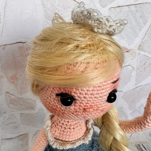 May include: A crocheted doll with blonde hair wearing a blue dress and a white crown. The doll has black eyes and a pink face.