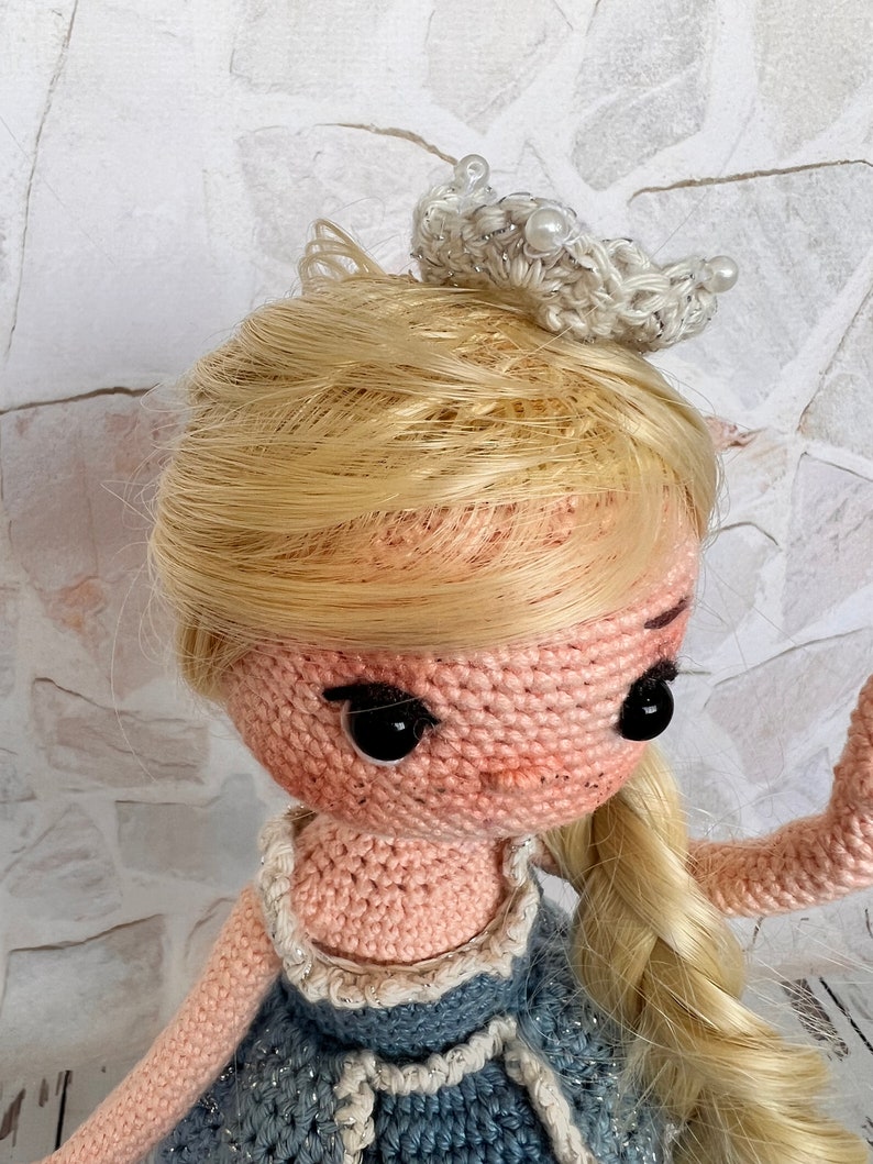 May include: A crocheted doll with blonde hair wearing a blue dress and a white crown. The doll has black eyes and a pink face.