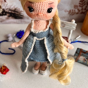 May include: A crocheted doll with long blonde hair, wearing a blue dress with white trim and white shoes.