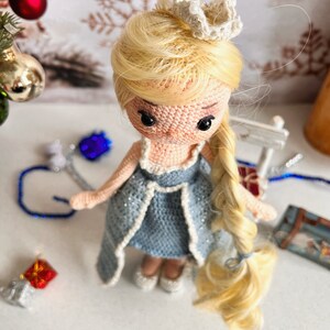 May include: A crocheted doll with blonde hair, wearing a blue dress and a white crown. The doll is standing on a white surface.