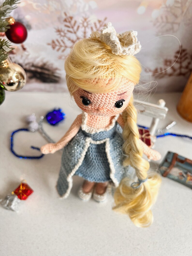 May include: A crocheted doll with blonde hair, wearing a blue dress and a white crown. The doll is standing on a white surface.