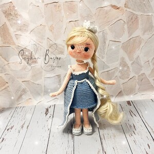 May include: A crocheted doll with blonde hair wearing a blue dress and a crown. The doll is standing on a wooden surface.